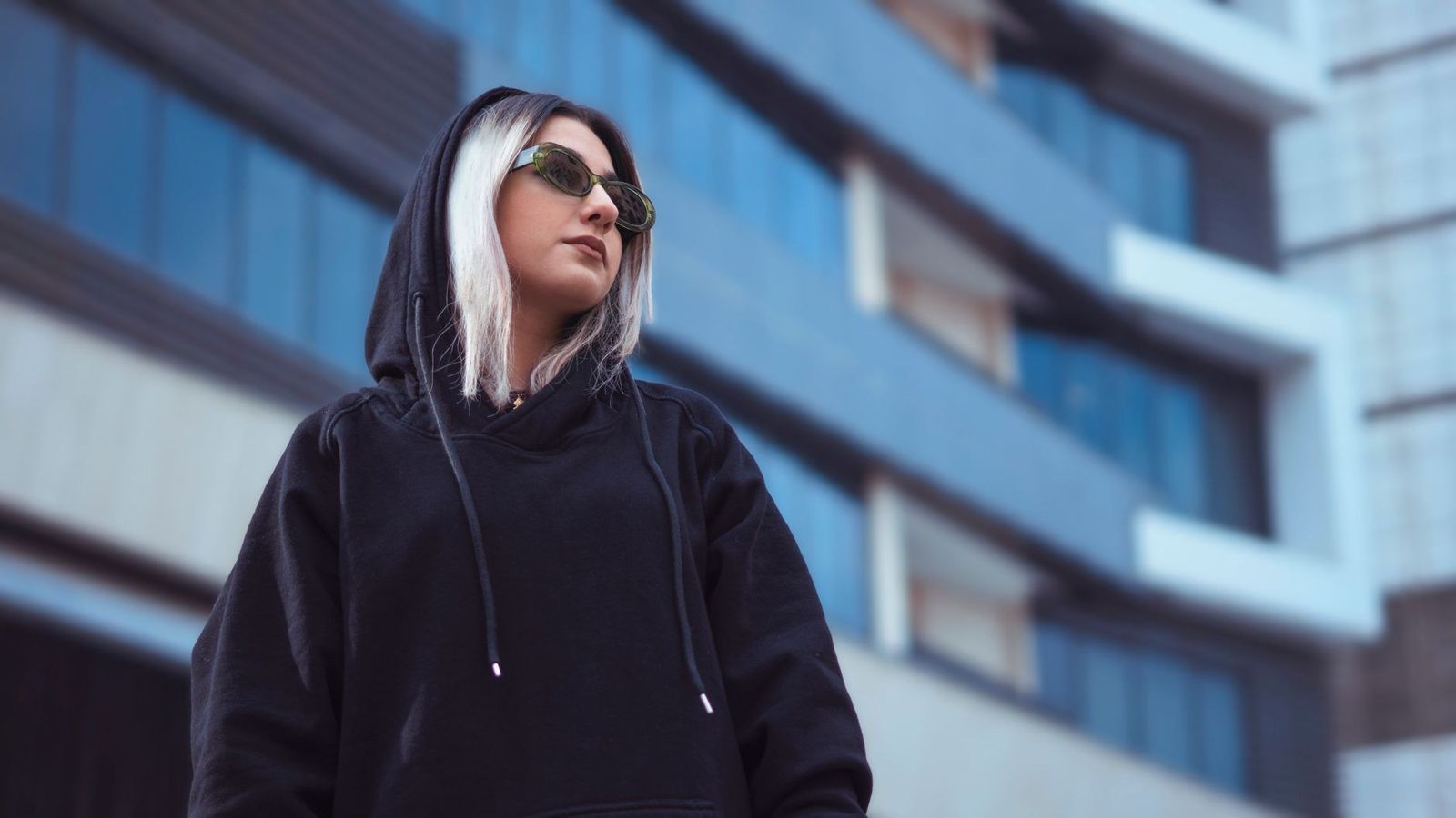 Woman in black hoodie and sunglasses stands confidently in front of modern building.